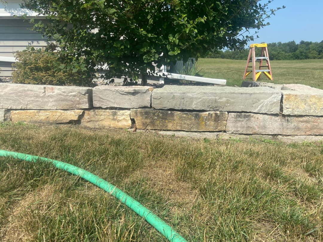 Stone landscaping wall with green garden hose and ladder in grassy yard.