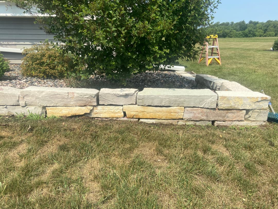 Stone retaining wall in garden with yellow and pink accents, surrounded by grass and plants.