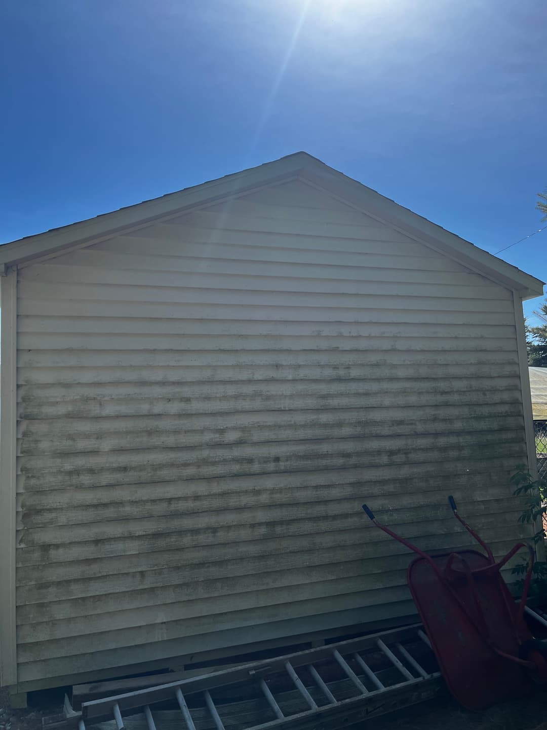 Weathered storage shed with visible stains and a wheelbarrow in the foreground under bright sunlight.