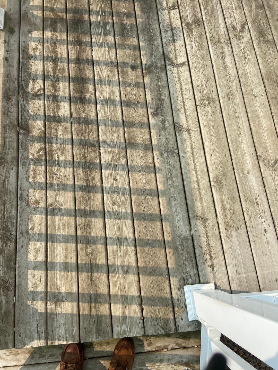 Sunlit wooden deck with shadows cast from a railing, featuring a pair of brown shoes.
