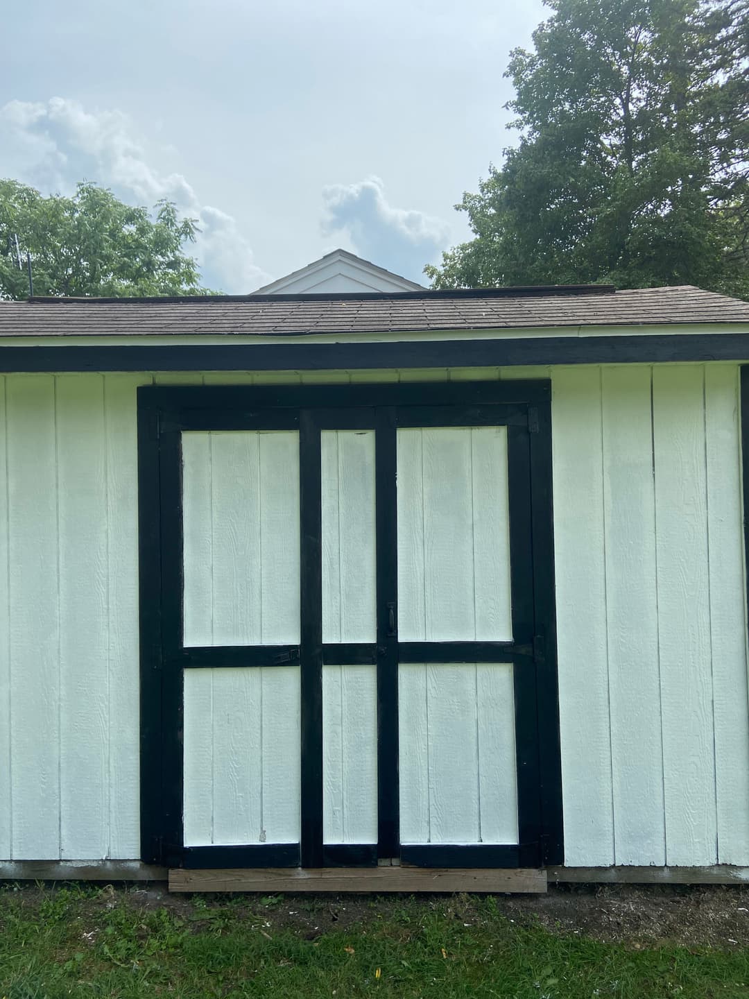 White shed with black doors and a slanted roof, surrounded by greenery.