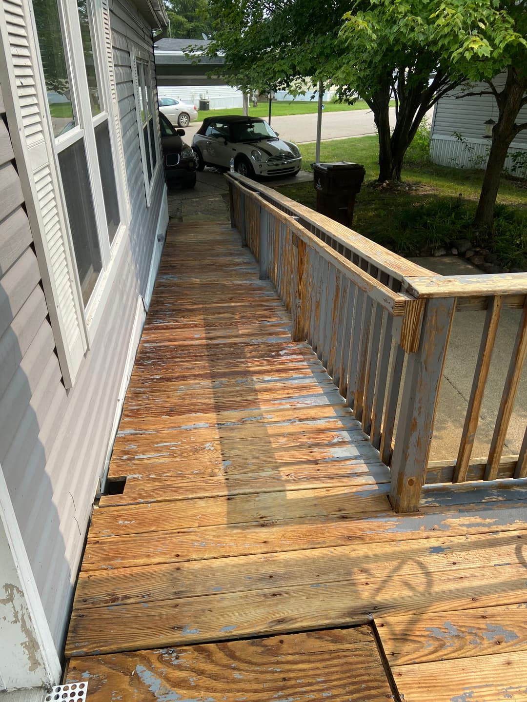 Wooden walkway with a railing leading to a house, surrounded by greenery and cars.