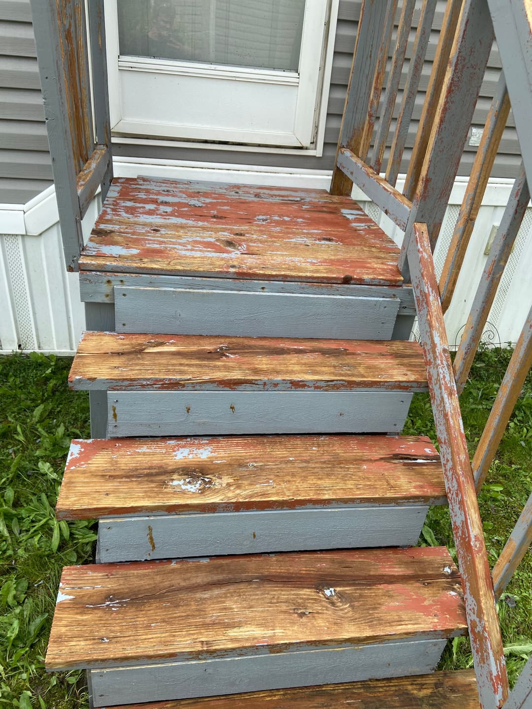 Worn wooden stairs with peeling paint leading to a door, surrounded by green grass.