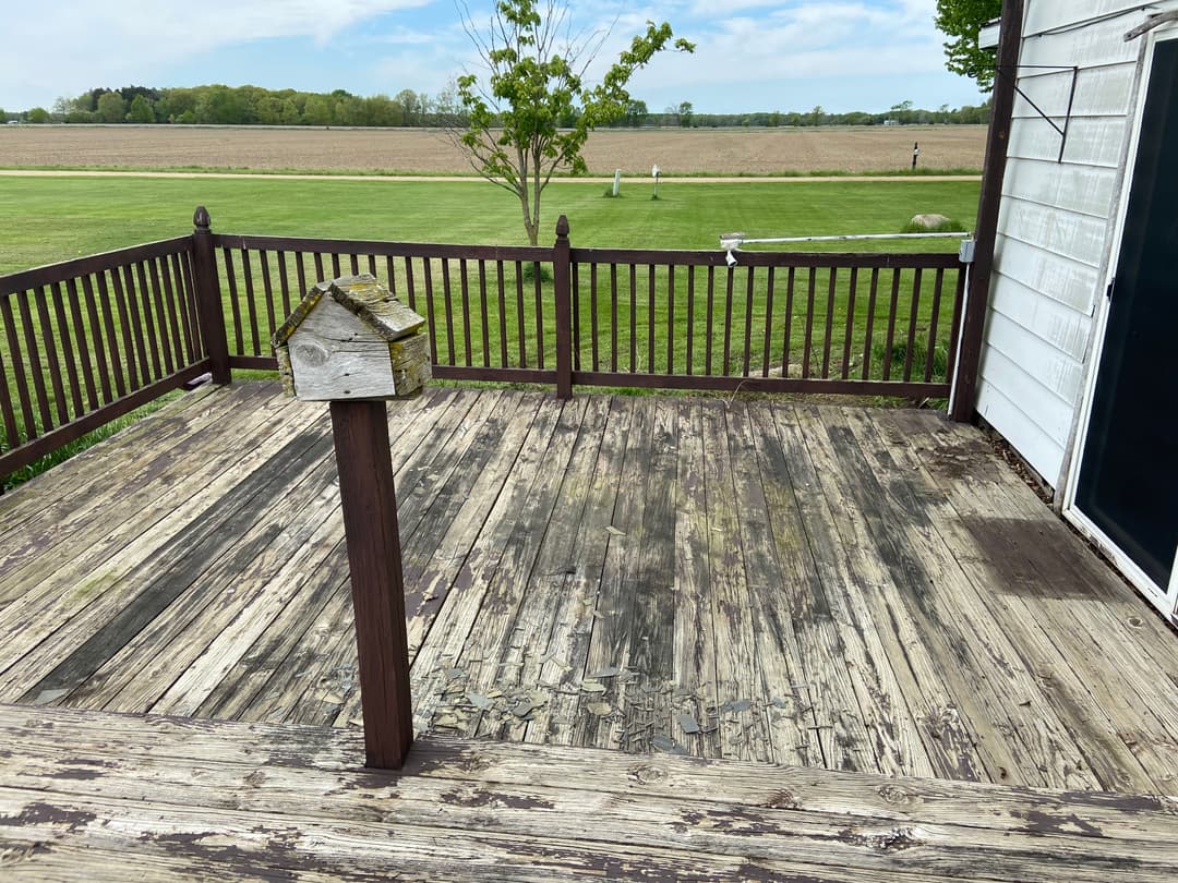 Weathered wooden deck with a birdhouse, overlooking a grassy field and distant trees.