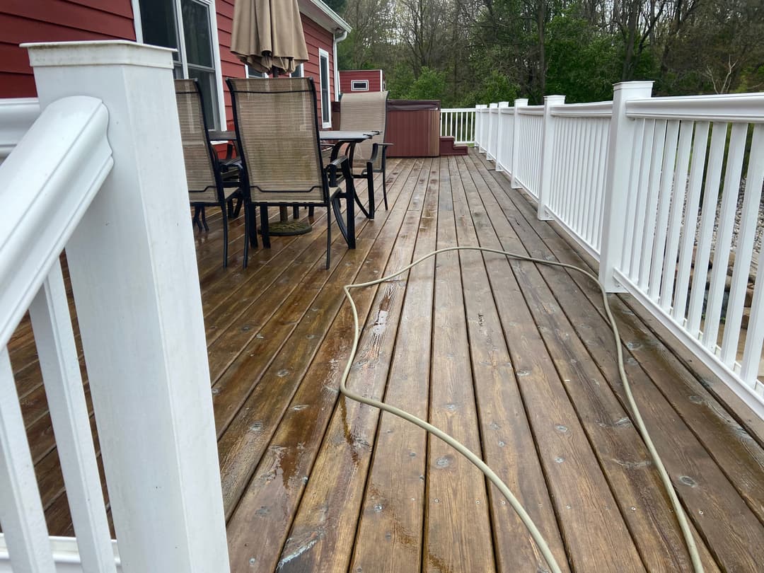 Deck with wooden flooring, patio furniture, and a hose, surrounded by greenery and red house.