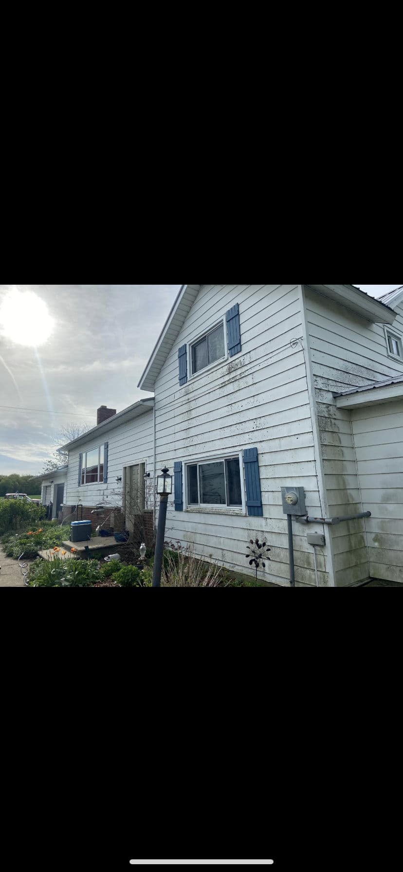 Two-story house with blue shutters, overgrown garden, and sunlit sky in the background.