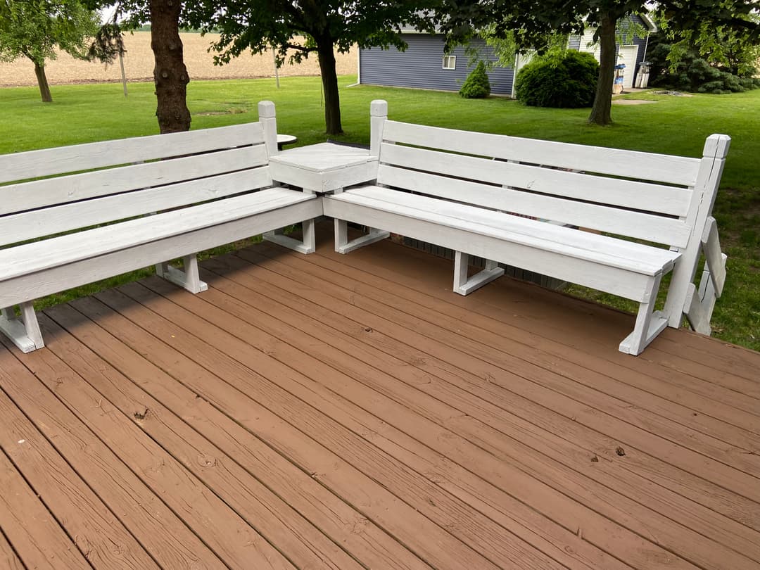 White wooden corner benches on a brown deck surrounded by a green lawn and trees.