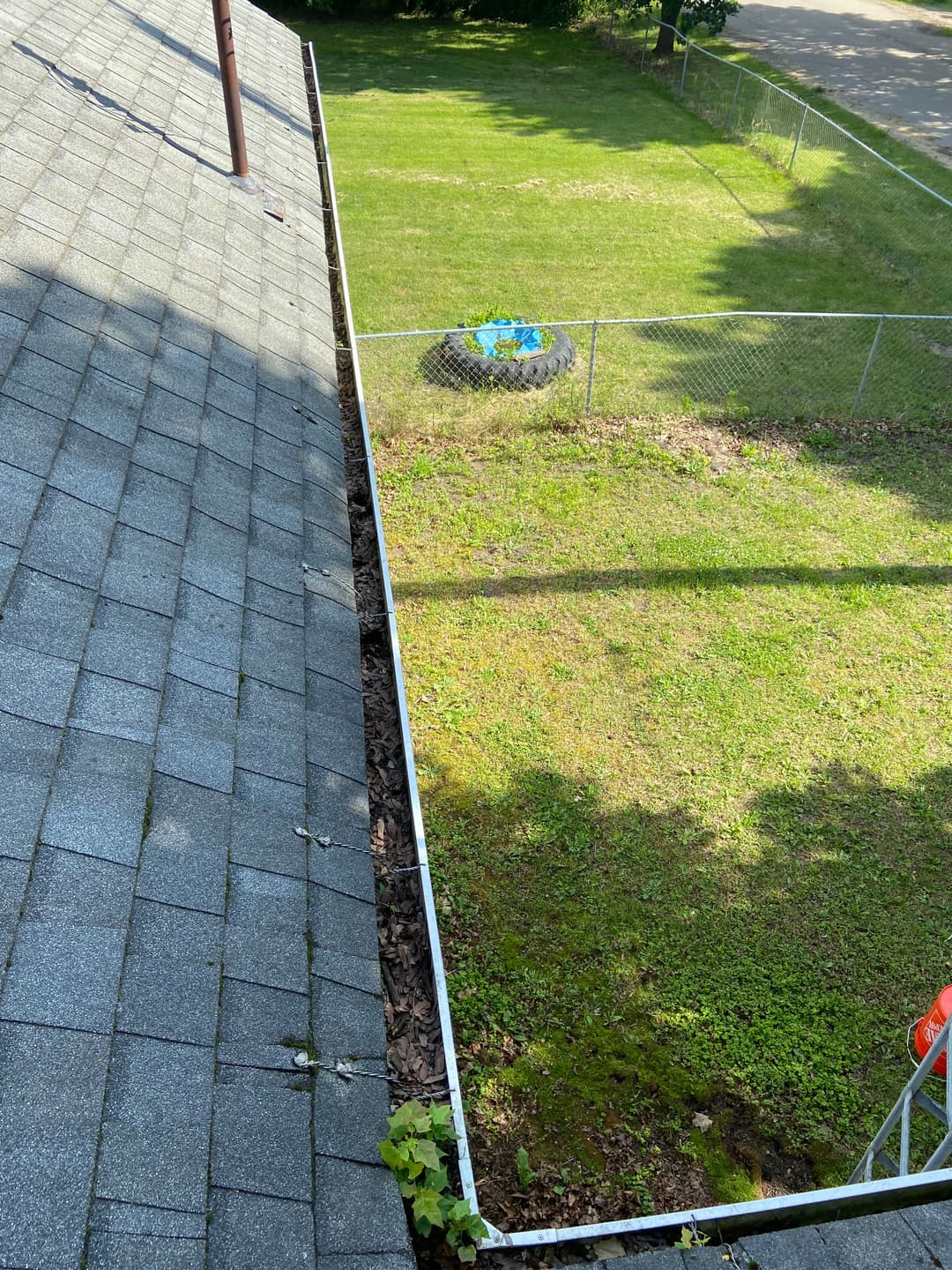 Aerial view of a roof edge with moss, showing a grassy yard and a tire in a fenced area.