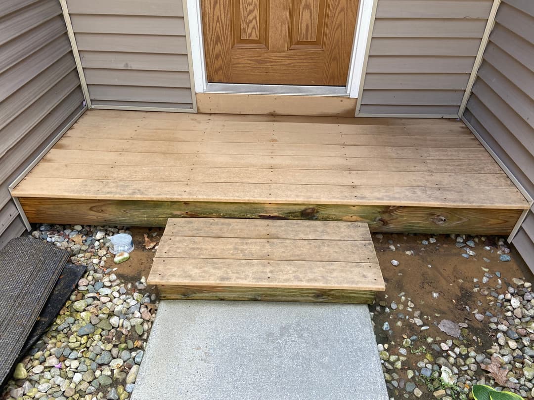 Wooden entryway porch with steps and gravel pathway, leading to a brown front door.