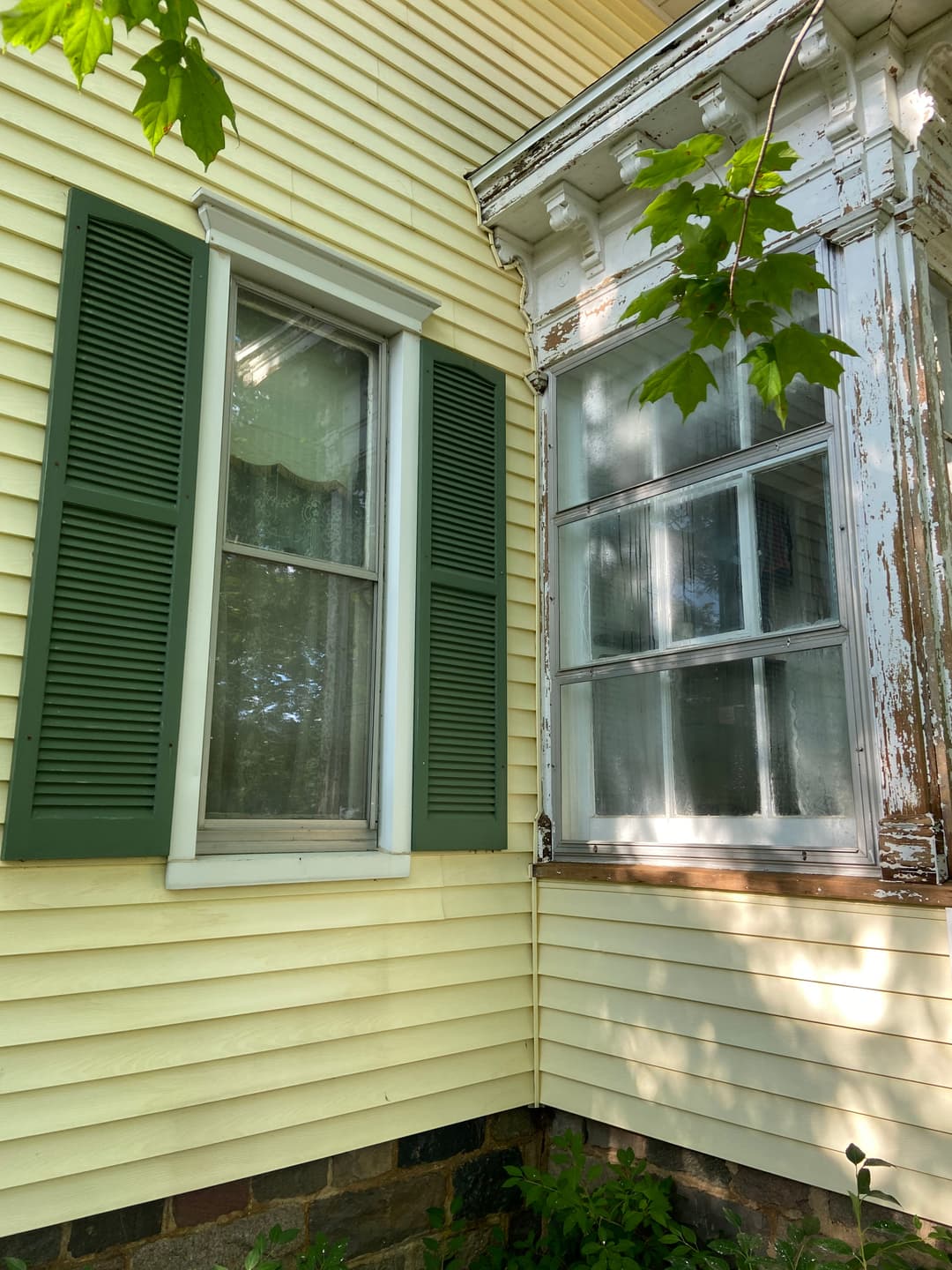 Close-up of a yellow house featuring green shutters and an old bay window with peeling paint.