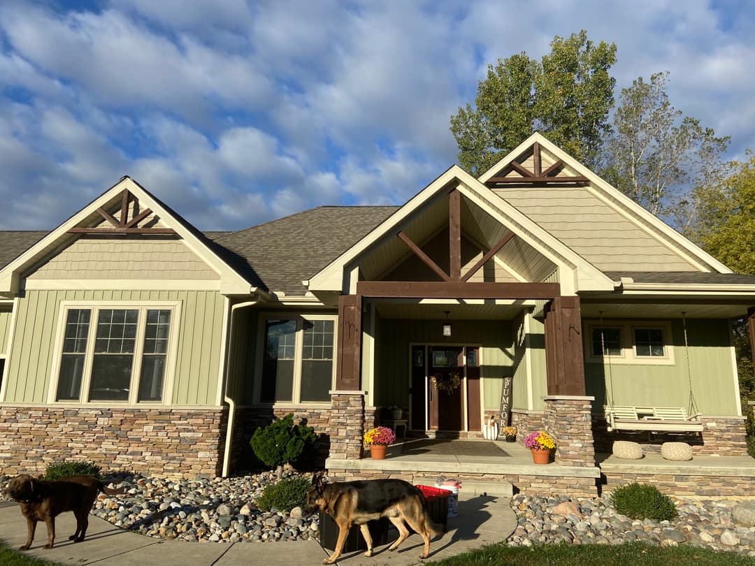 Modern green house with stone accents, large porch, and two dogs in front yard.