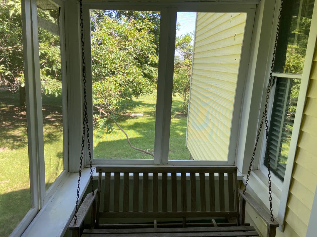 Screened porch with a wooden swing bench overlooking a green backyard and trees.