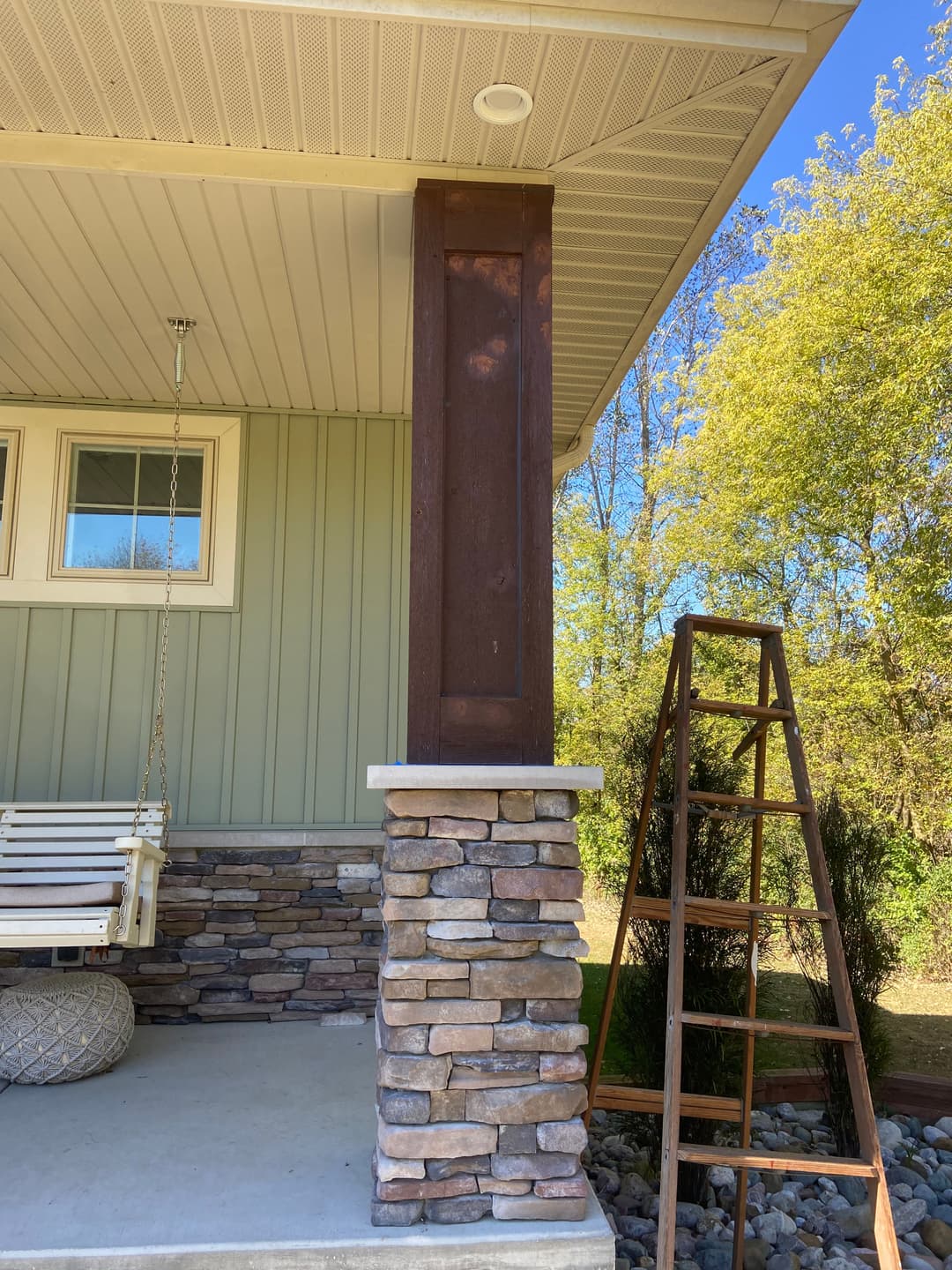 Wooden column on a front porch, featuring stone base and a ladder nearby, with greenery in the background.