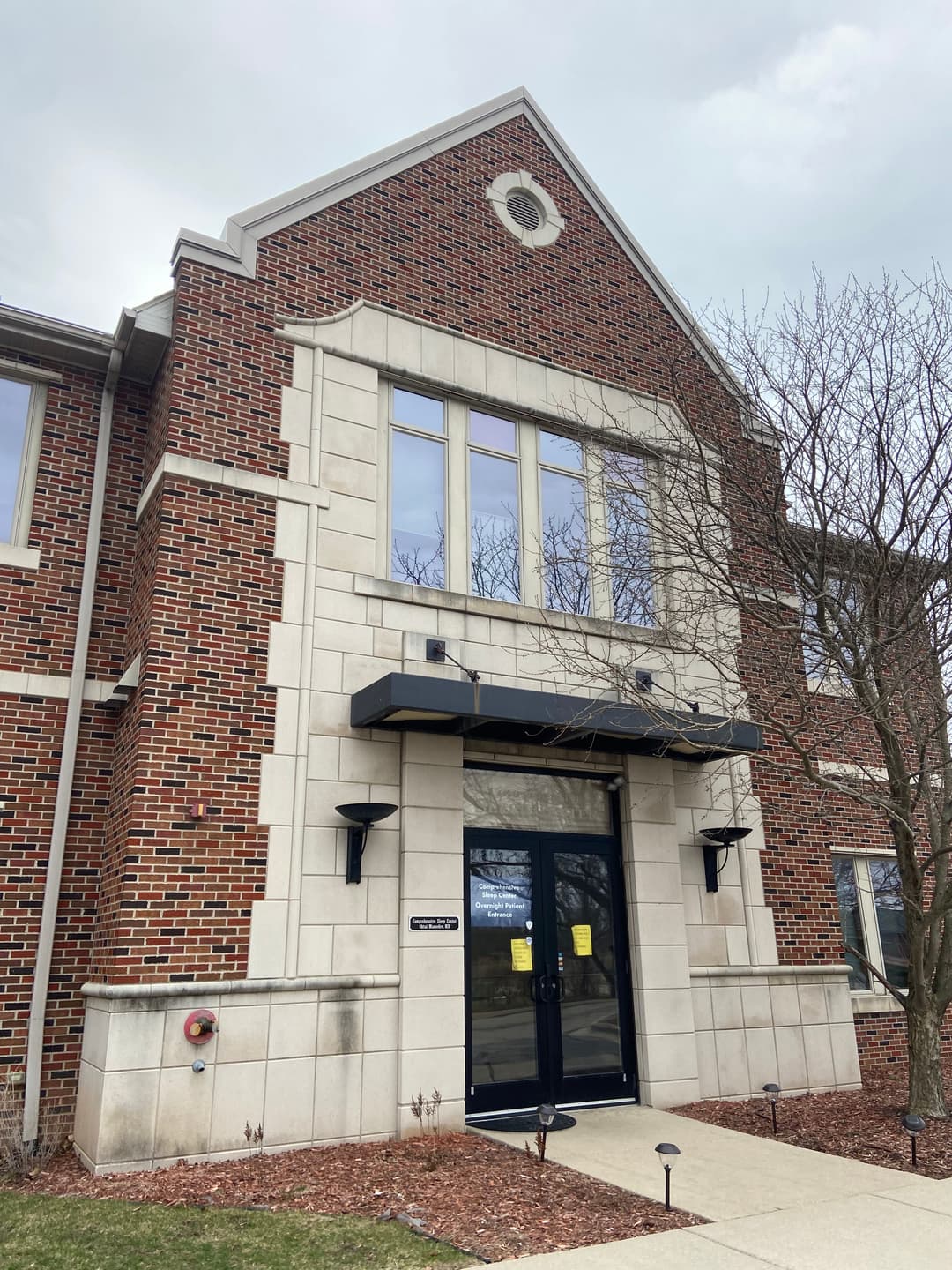 Brick building entrance with large windows and black door canopy on a cloudy day.