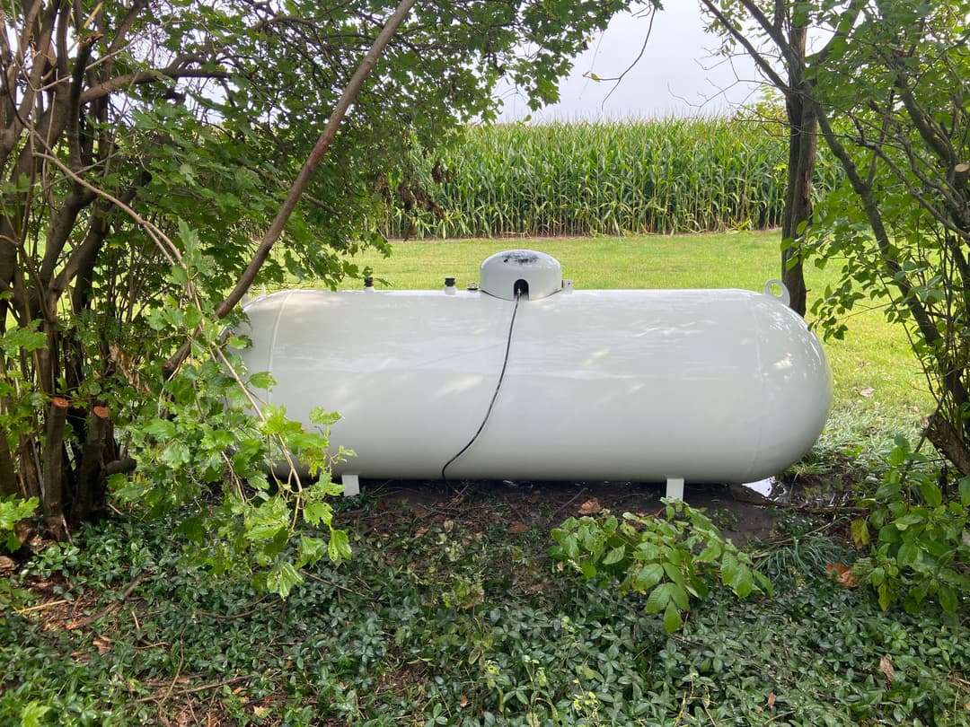 White propane tank partially hidden by trees in a grassy area near a cornfield.