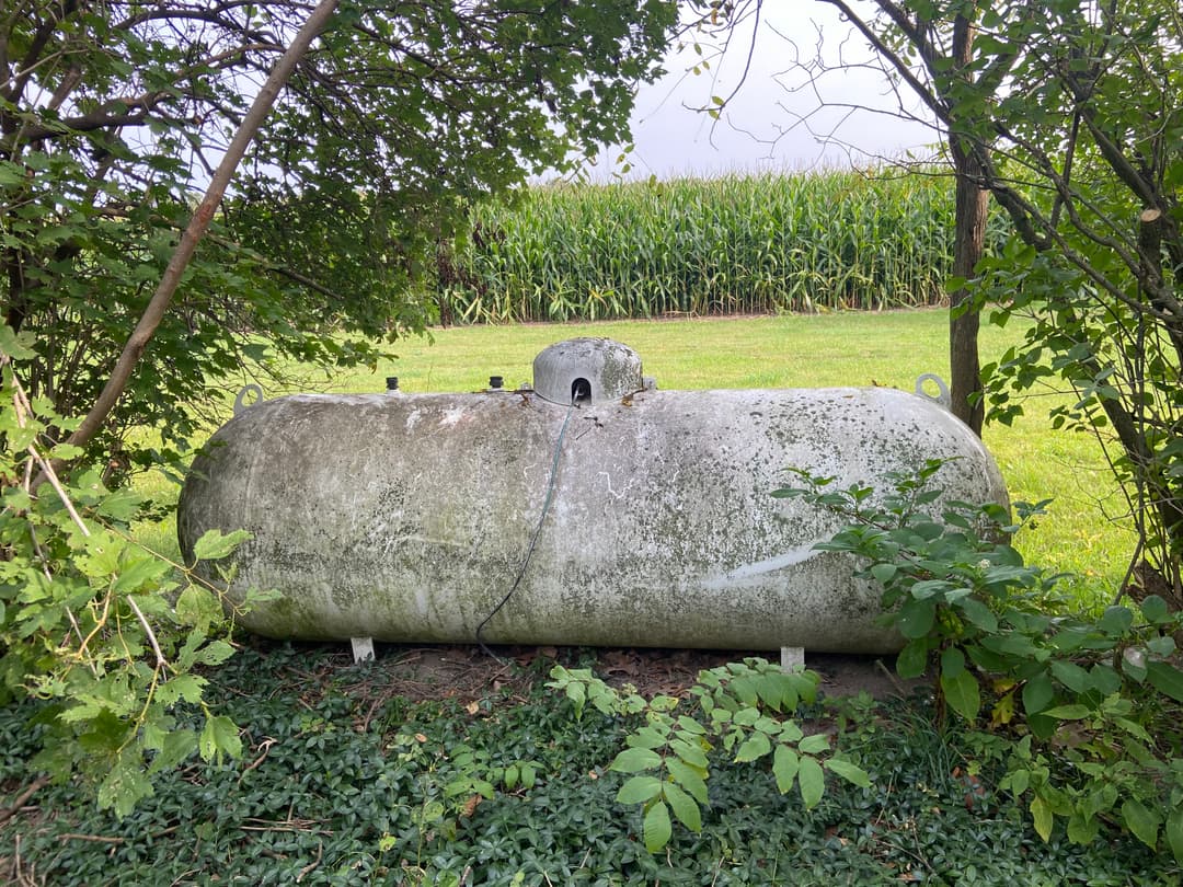 Weathered propane tank surrounded by greenery near a cornfield.