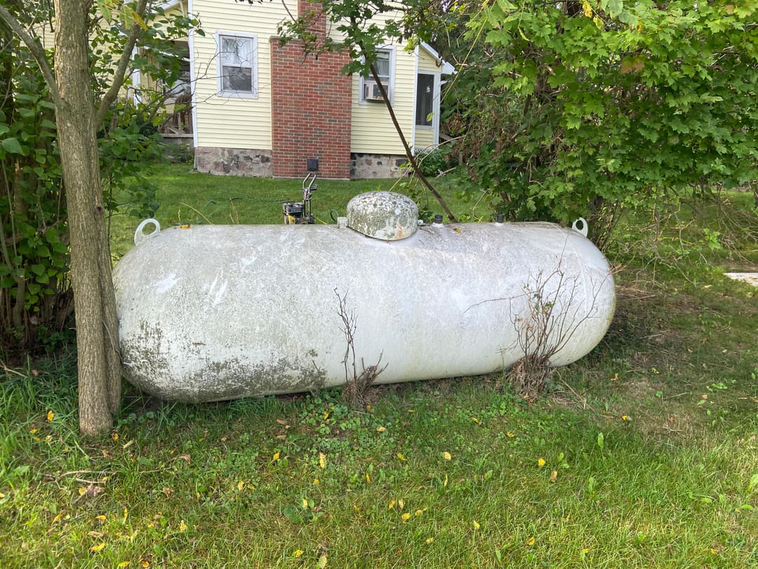 Old propane tank beside a house, partially overgrown with grass and nearby trees.
