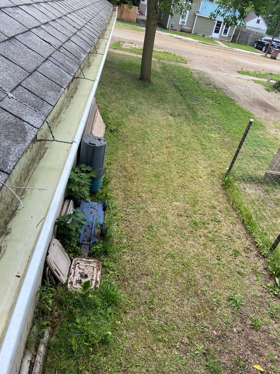 View of a grassy yard with trash bins next to a house, surrounded by trees and fence.