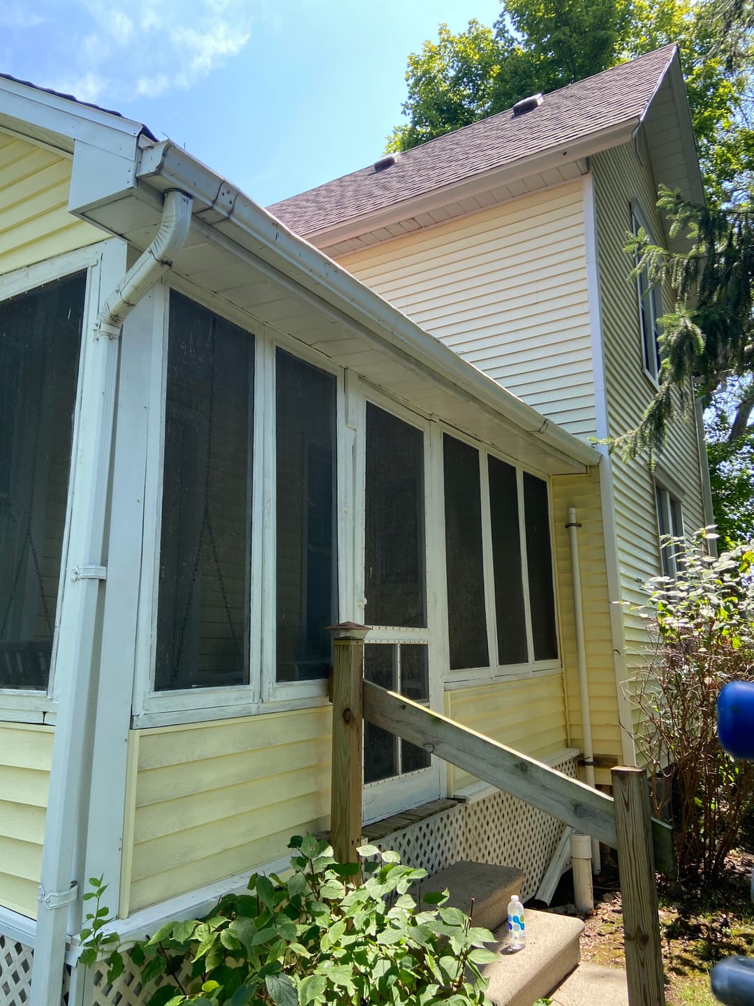 Screened-in porch on a yellow house with green foliage and a sunny sky in the background.