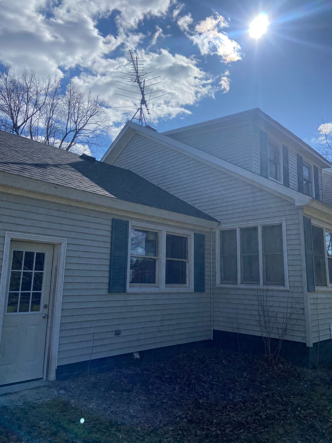 Side view of a gray house with window shutters and an antenna under a sunny sky.