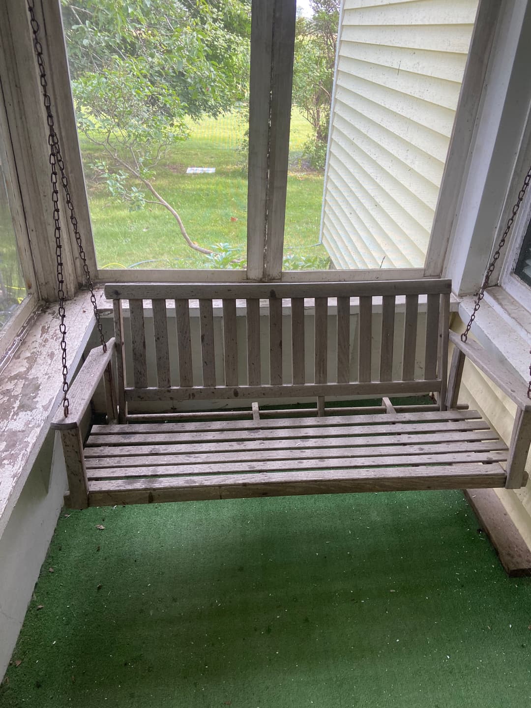 Weathered wooden swing on a screened porch with green carpet and natural light.