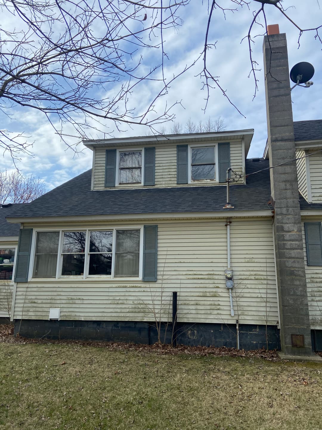 Two-story house with a gray roof, chimney, and blue shutters under a cloudy sky.