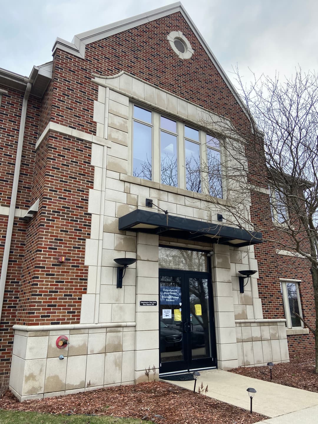 Modern brick building entrance with large windows, a black awning, and landscaped surroundings.